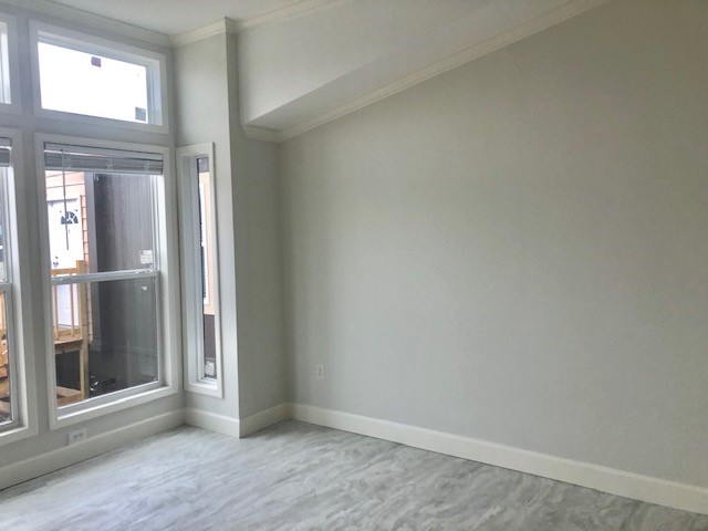 Empty room with light gray walls and a sloping ceiling. Large windows on the left side allow natural light, brightening the gray marble floor and white trim.