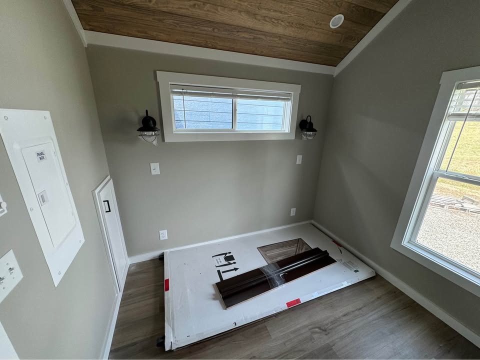 A small, empty room with light gray walls and wood flooring. A large window brightens the space, and a wood ceiling adds warmth. Two wall-mounted lamps are beside a narrow window.