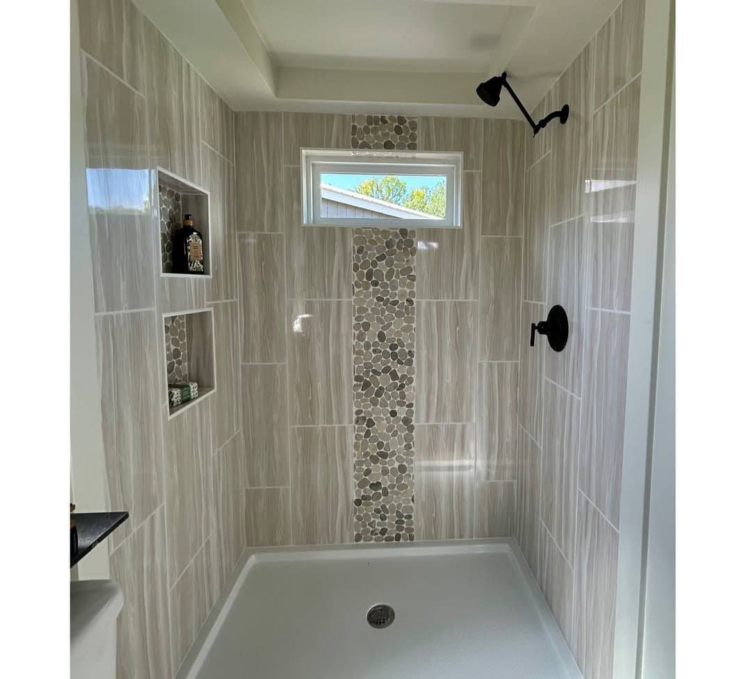 Modern shower with beige tiles, pebble accent strip, and inset shelves holding toiletries. A small window provides natural light, creating a calm ambiance.
