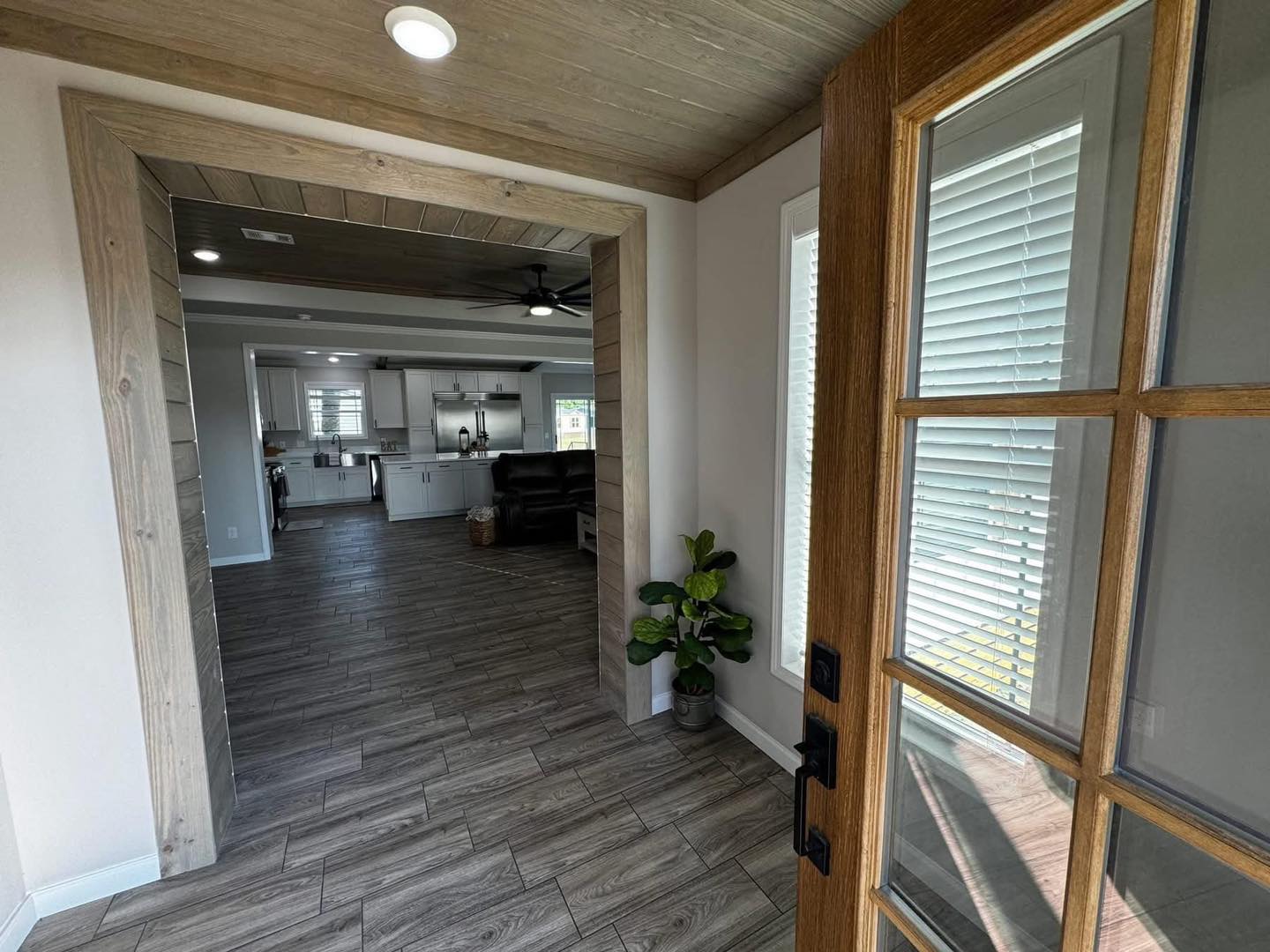 View of a modern, cozy home interior from the open door. Features wood floors, a potted plant, and a bright kitchen with white cabinets in the background.