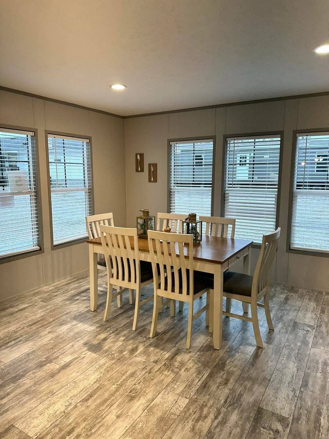 A cozy dining room with a wooden table and six chairs on a light wooden floor. Large windows with blinds allow natural light, creating an inviting atmosphere.