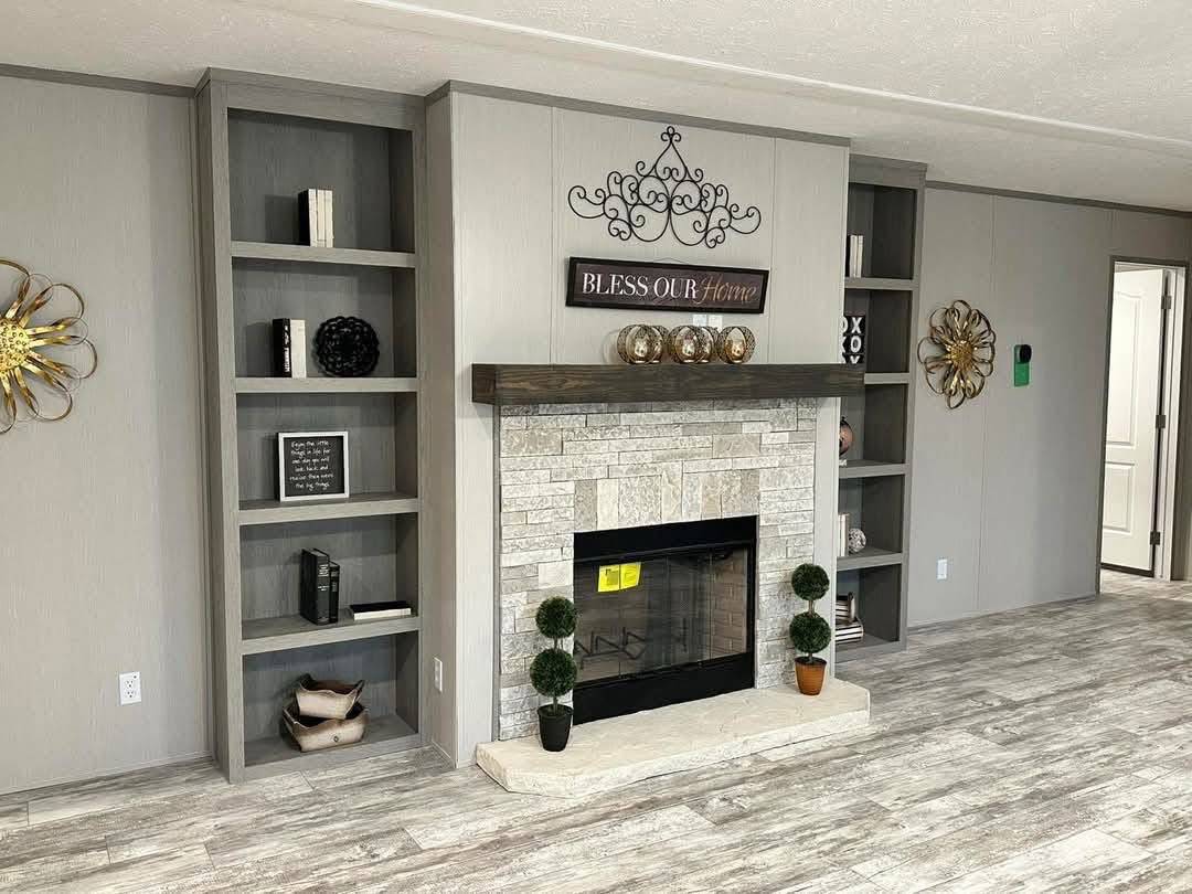 Cozy living room with a stone fireplace, decorative mantel items, and greenery. Built-in shelves hold books and ornaments; "Bless Our Home" sign above.