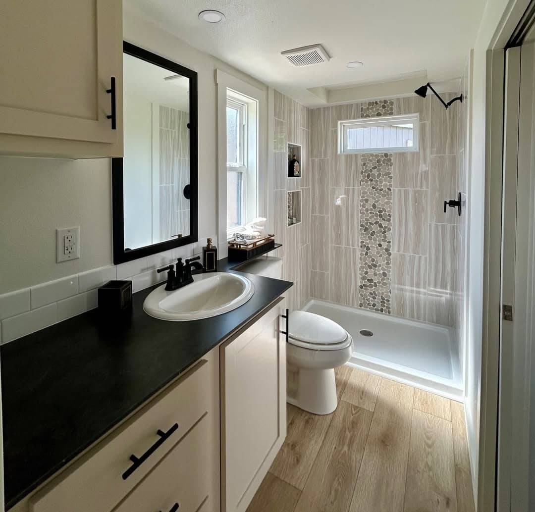A modern bathroom with light wood flooring, a black-counter vanity, and white cabinets. It features a glass shower with pebble tile accents and natural light from the window.