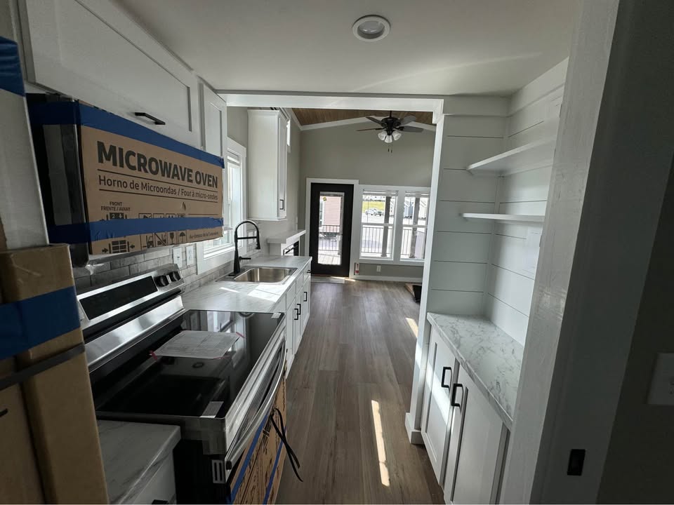 Narrow modern kitchen with white cabinets, stainless steel appliances, and light wood flooring. Sunlit living area visible through door, creating an airy feel.