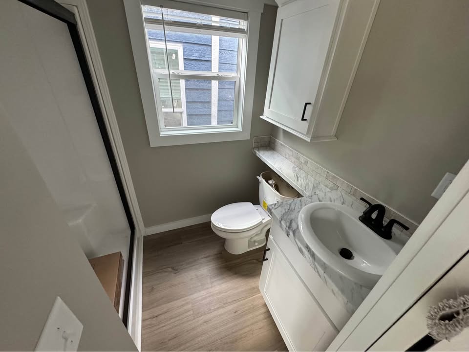 Compact bathroom with a small shower, toilet, and sink. White cabinetry, marble countertop, light gray walls, and wood floor create a clean, modern feel.