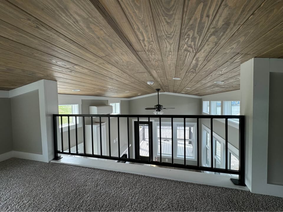 Loft interior with wood-paneled ceiling, modern railing, and carpeted floor. Large windows below allow natural light, creating an open, airy feel.
