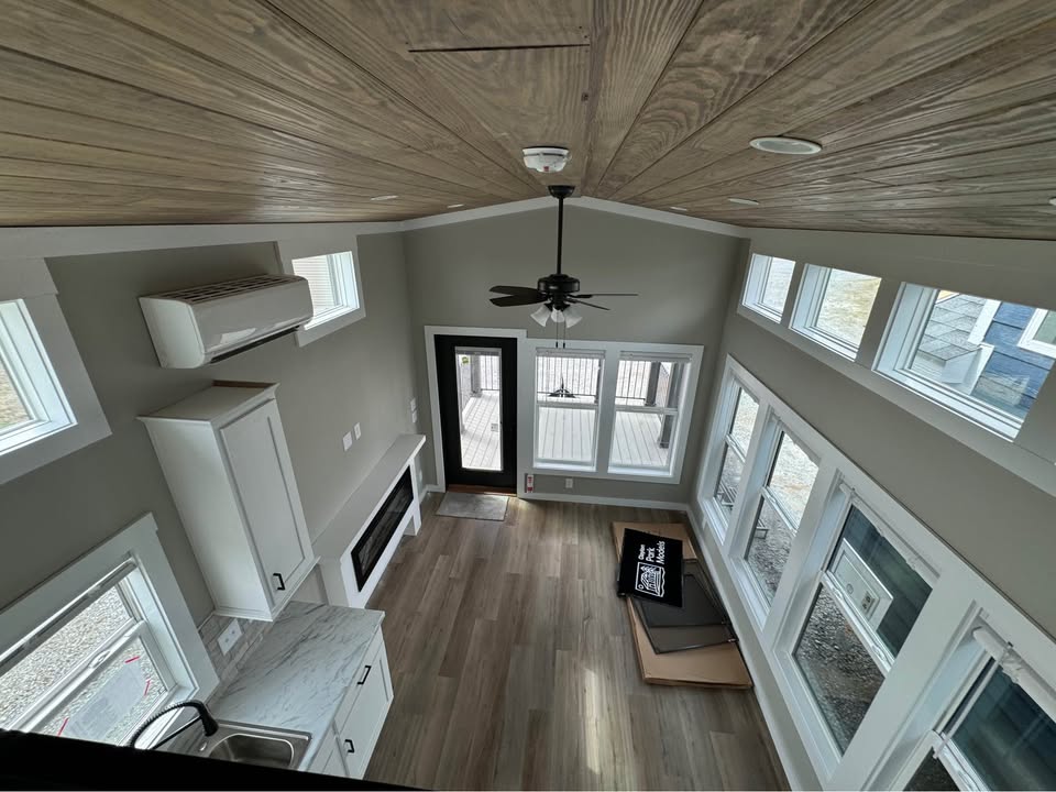 Spacious tiny home interior viewed from above, featuring wood flooring, a ceiling fan, large windows, a built-in fireplace, and air conditioner.