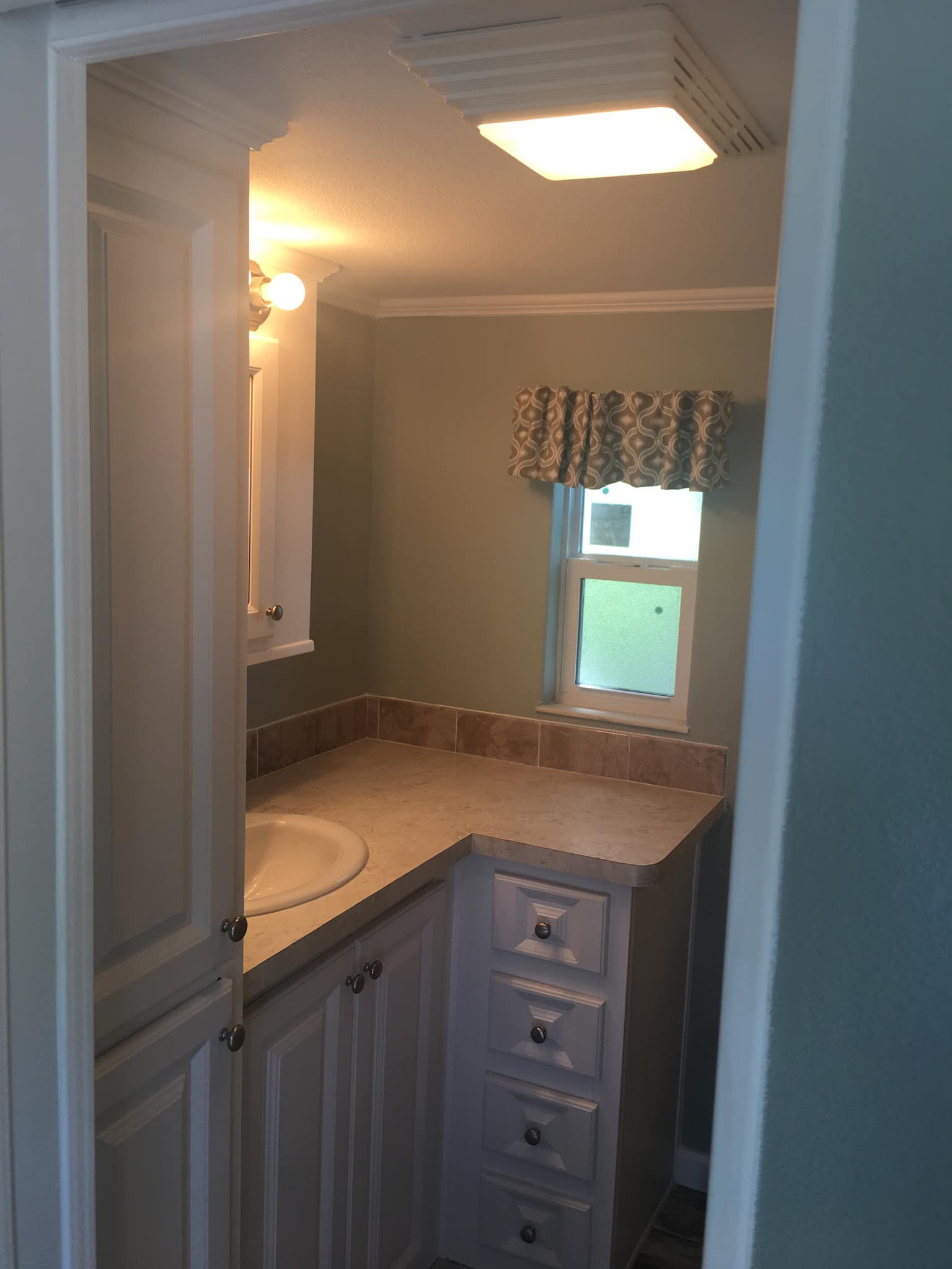 Compact bathroom with white cabinets and a light beige countertop. Soft light illuminates the green walls. Small window with patterned curtain.