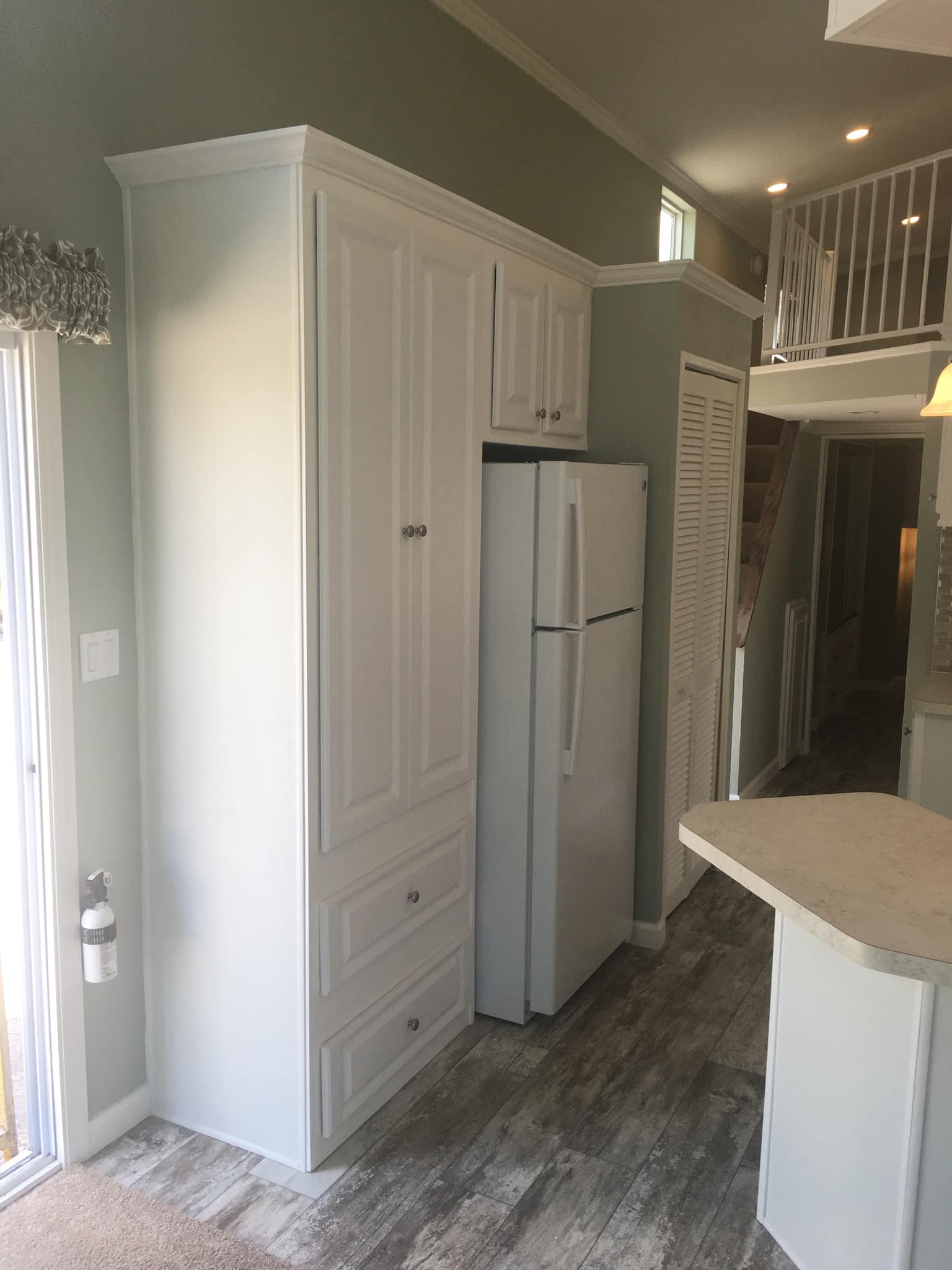 A cozy kitchen with a light tone, featuring white cabinets, a fridge, and wooden floors. A loft with a railing is visible, adding height and openness.