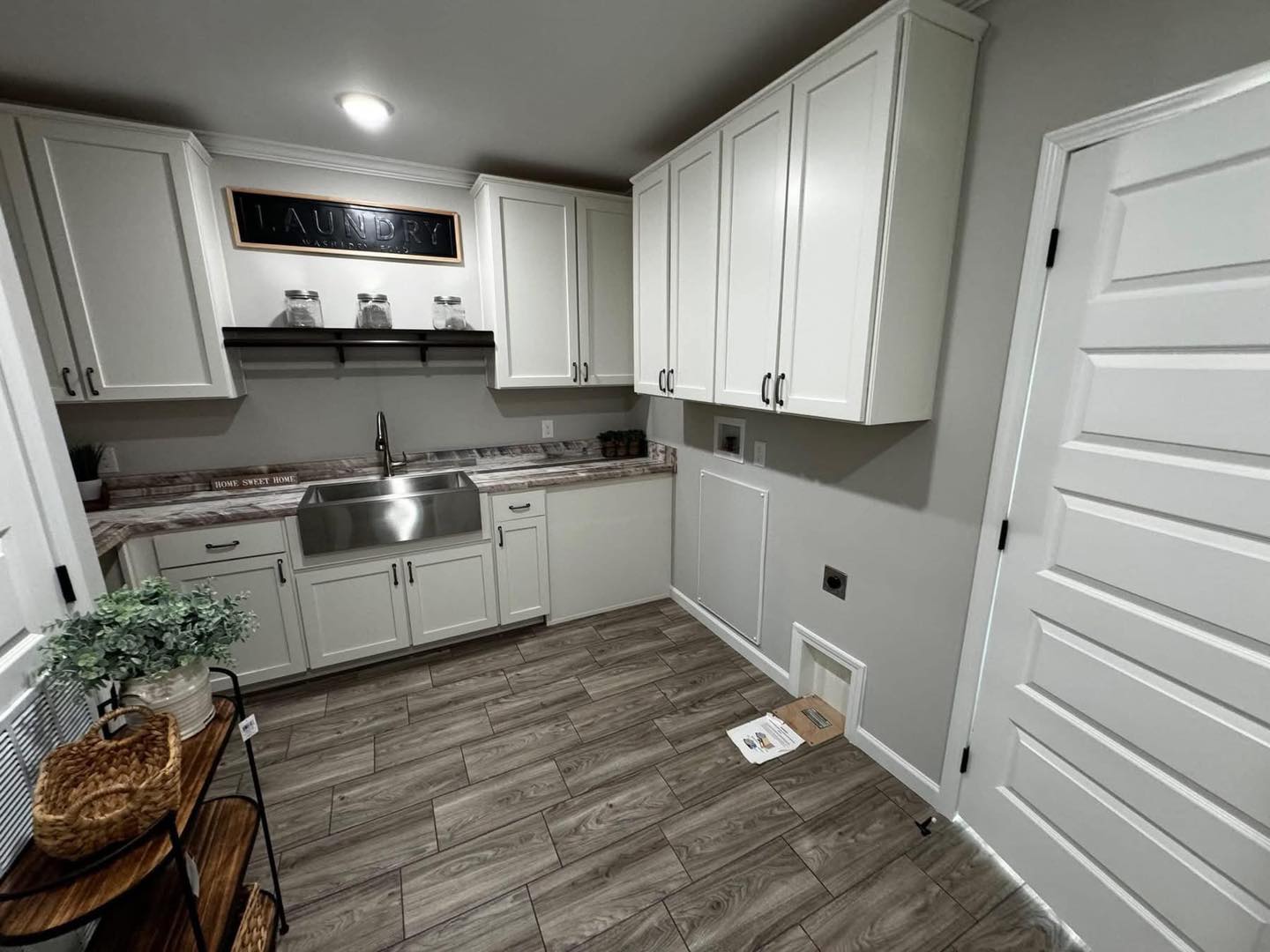 Modern laundry room with white cabinets, large stainless steel sink, and wood-patterned tile flooring. A "Laundry" sign adds charm above shelves.