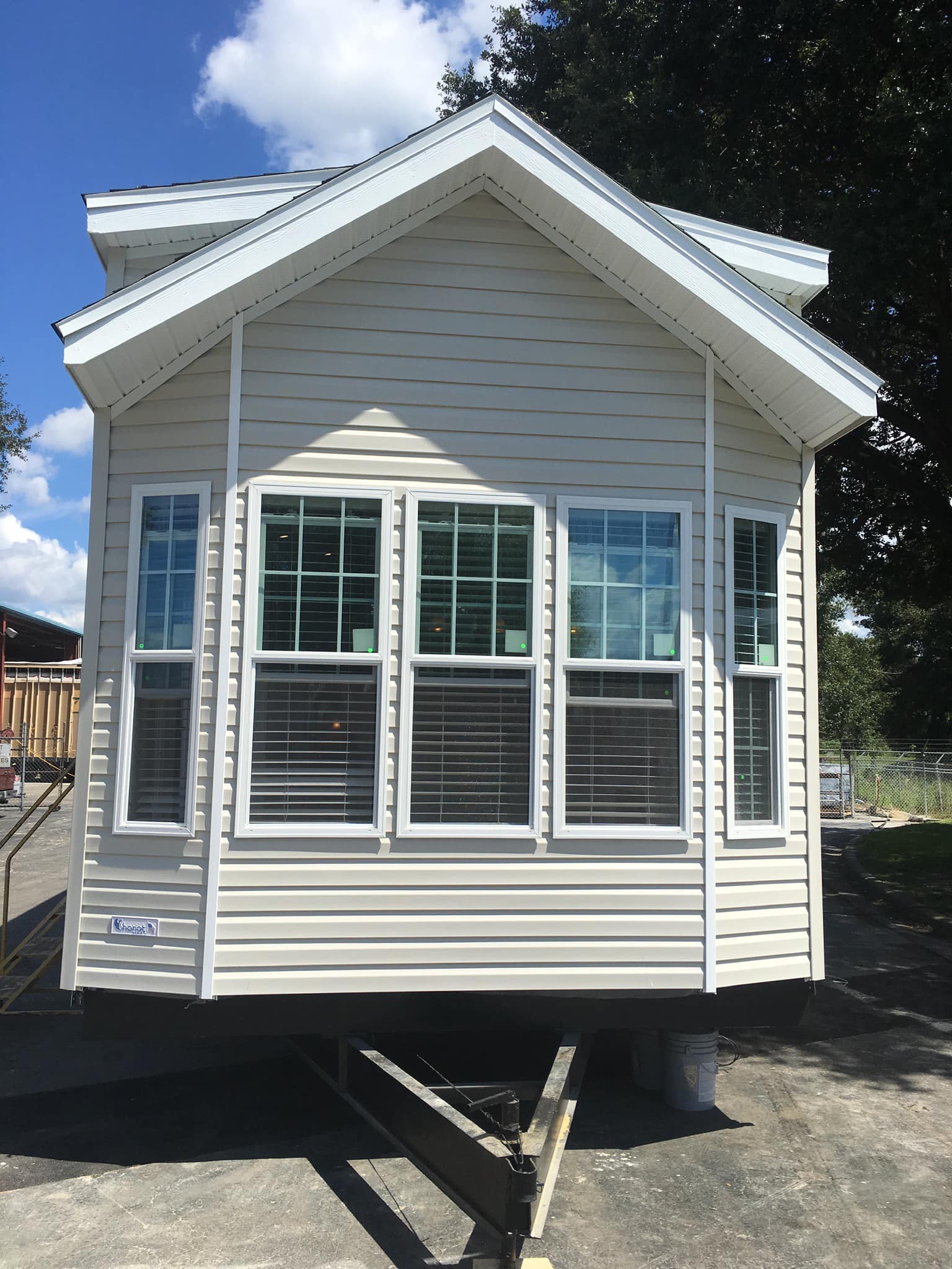 Small, beige park model home with large gabled roof and six tall windows in front. It's on a trailer frame under bright blue sky.
