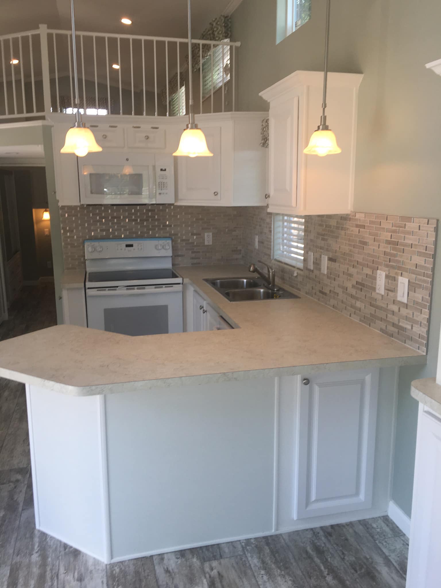 Bright kitchen with white cabinets, beige countertops, and a tiled backsplash. Three hanging lights illuminate the space. Wood-textured flooring adds warmth.