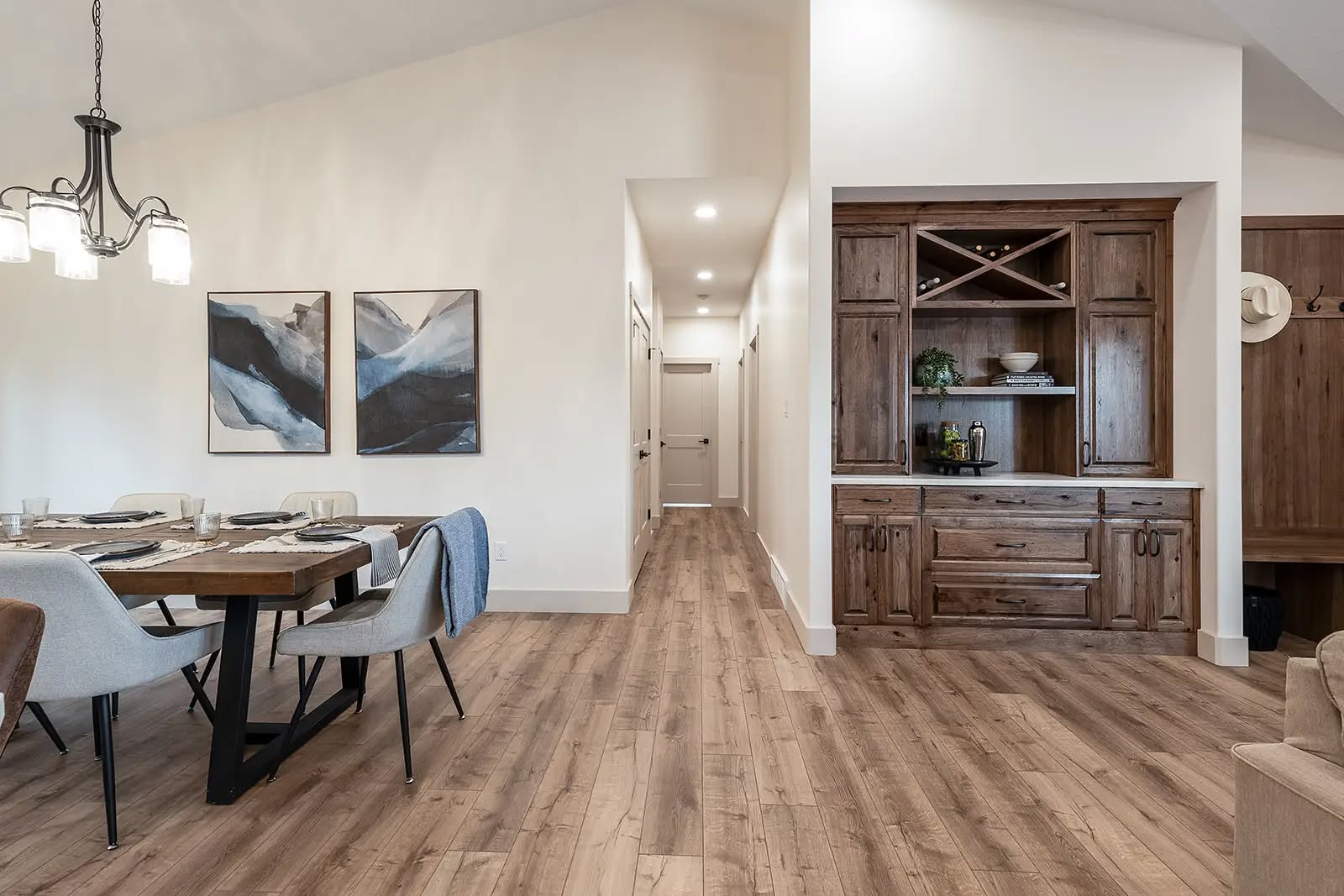 Spacious dining room with light wood floors, modern chandelier, abstract art, and wooden cabinets. Bright, airy feel with hallway and cozy decor.