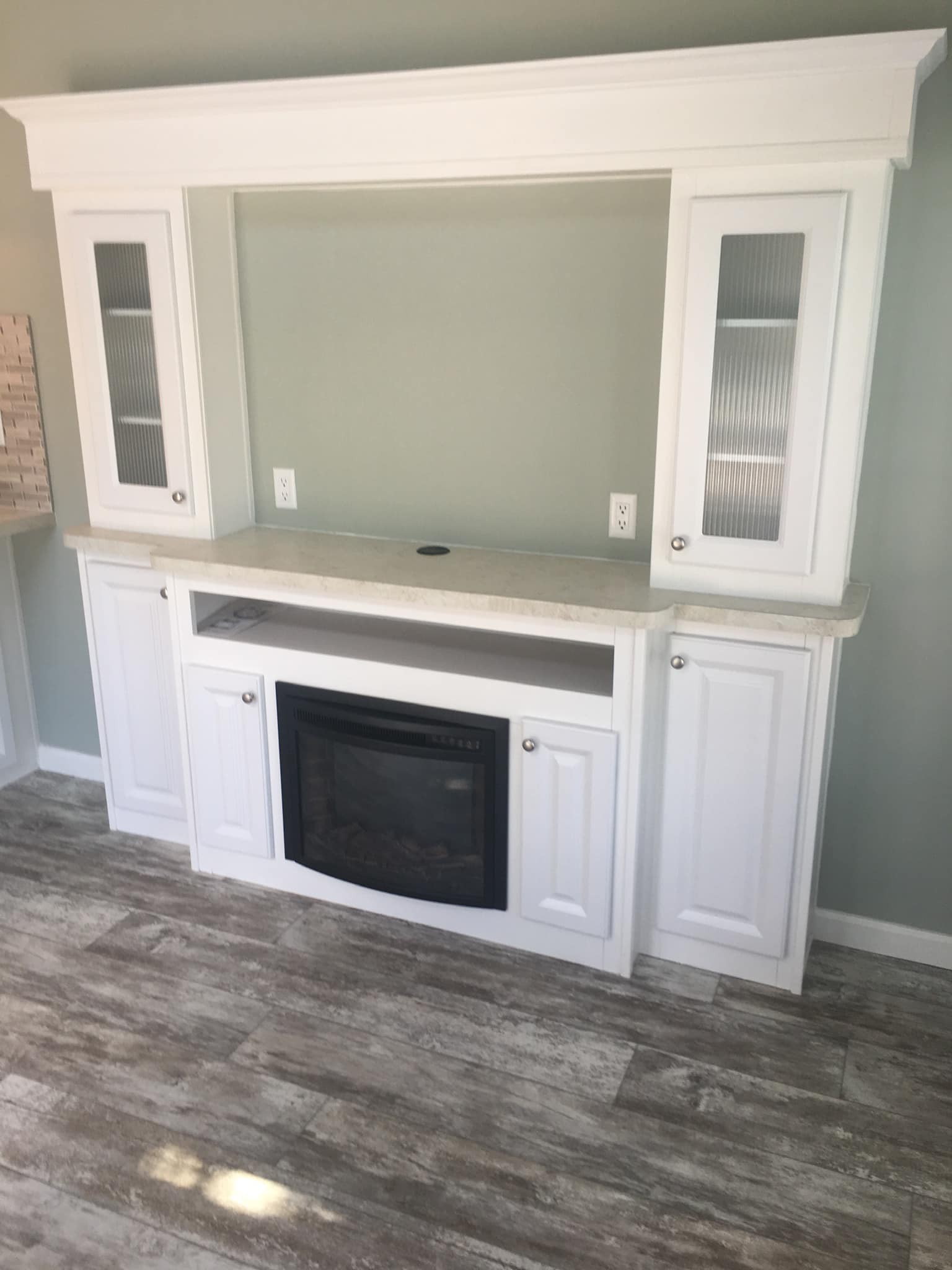 White entertainment unit with fluted glass cabinet doors and a central fireplace. It stands against a soft green wall on a wood-textured floor.