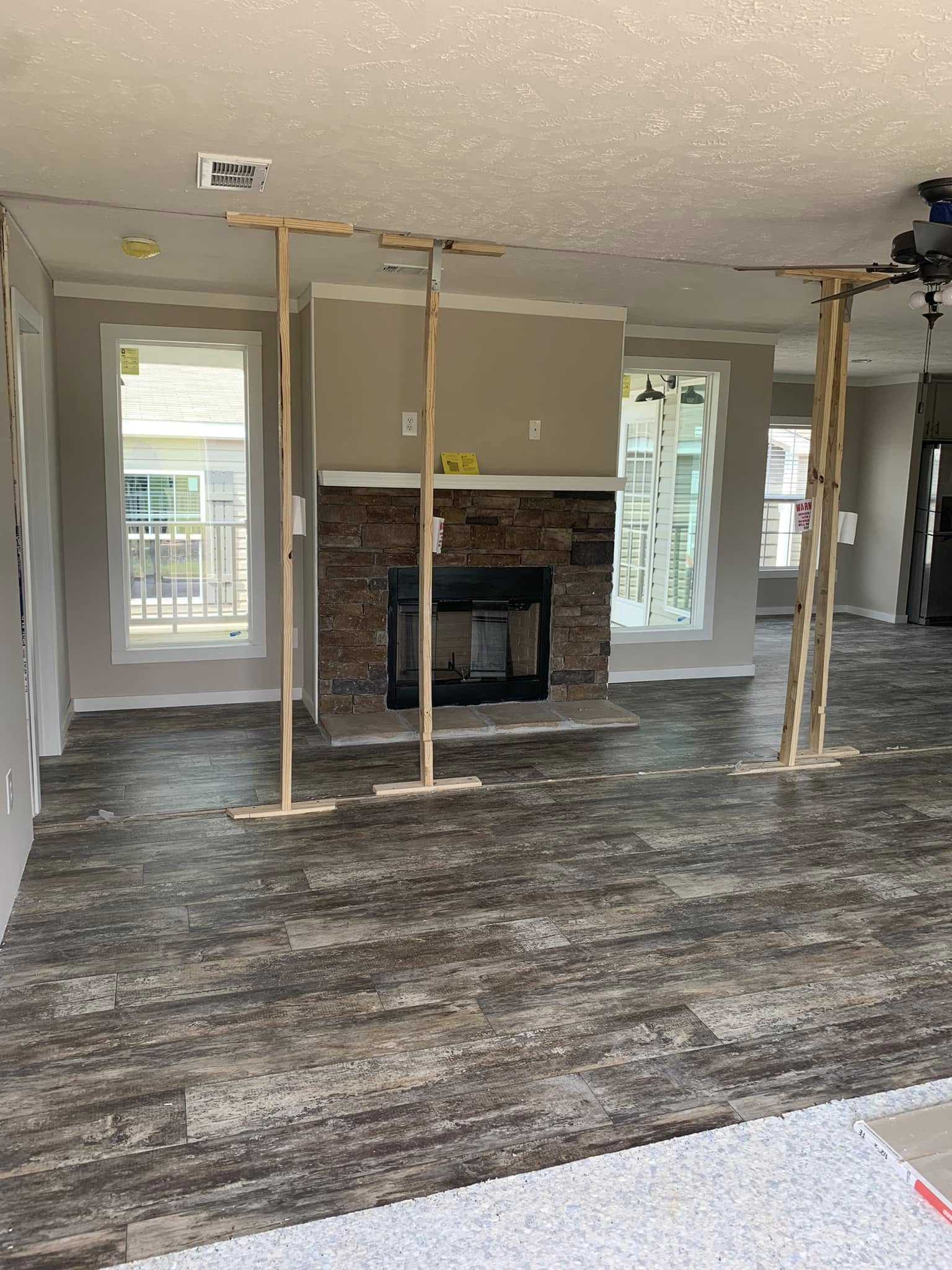 Living room under renovation with wooden supports holding the ceiling, a stone fireplace centered between tall windows, and dark wood flooring.