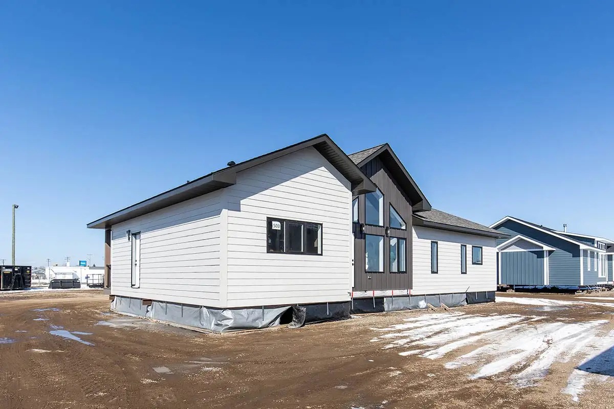 Modern prefabricated house with white siding and large windows under a clear blue sky. The house sits on a dirt lot with patches of snow.