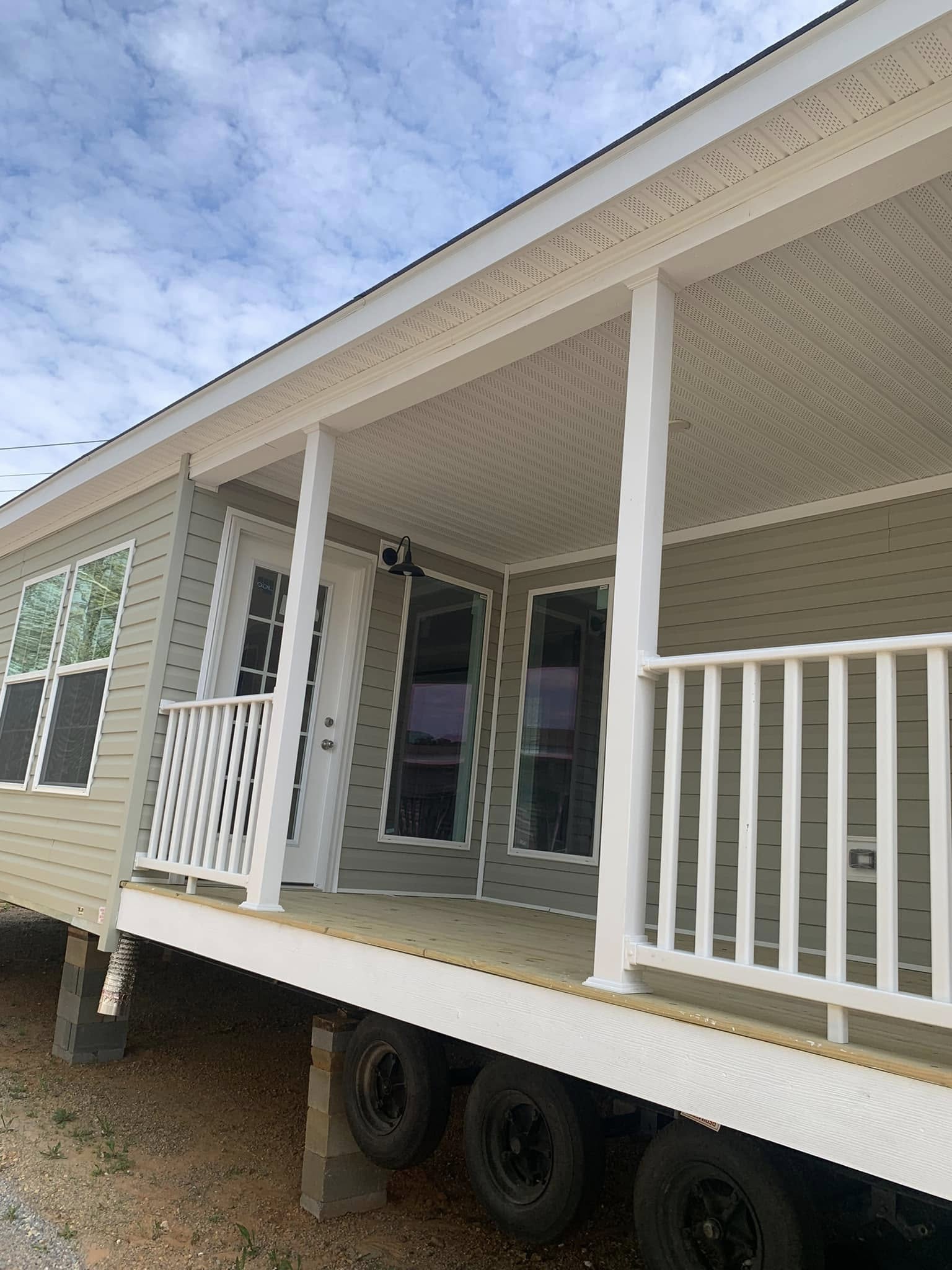 Cozy mobile home with green siding, white railing, and a porch. Large windows and a glass door face a cloudy sky. Calm and inviting atmosphere.