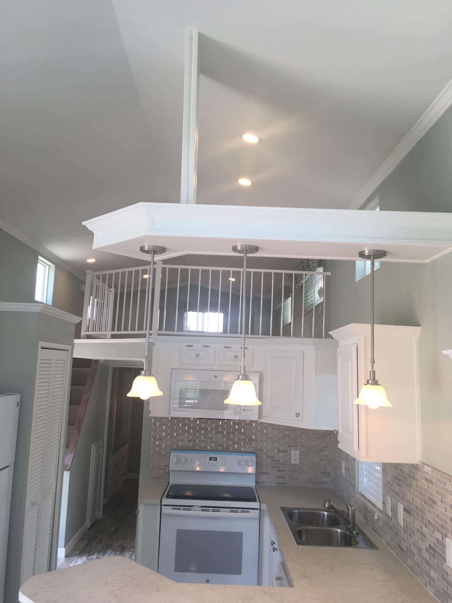 Modern kitchen in a tiny home featuring white cabinets, a tiled backsplash, and pendant lights. A loft with railing is visible above, enhancing the cozy feel.