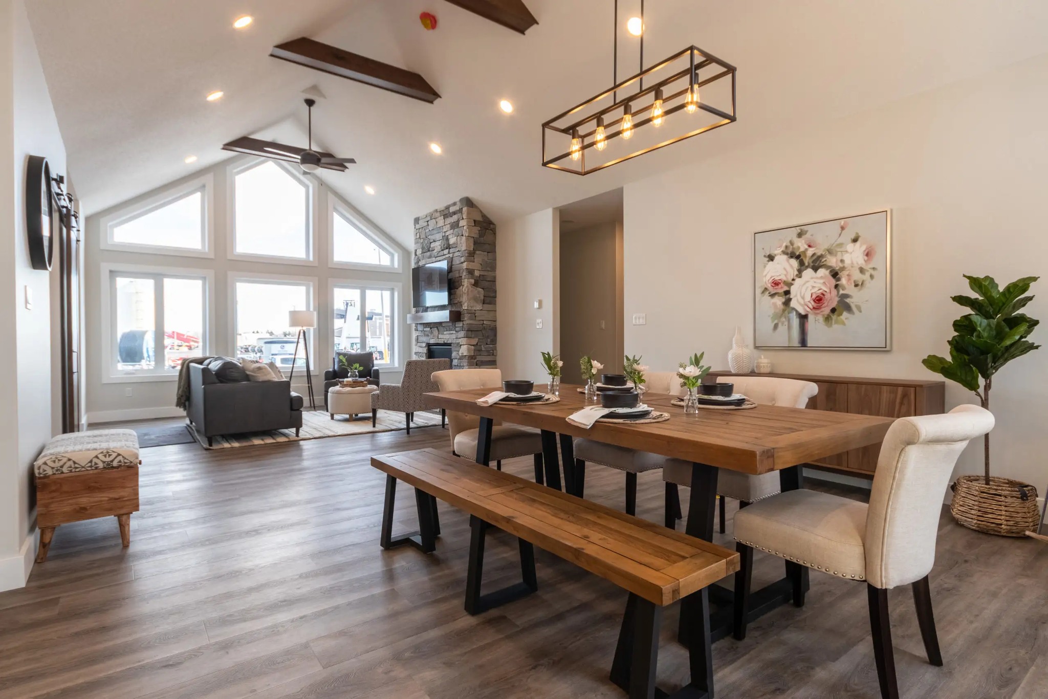 Spacious living room with vaulted ceiling and wood beams, featuring a wooden dining table, beige chairs, large windows, and stone fireplace. Cozy and elegant.