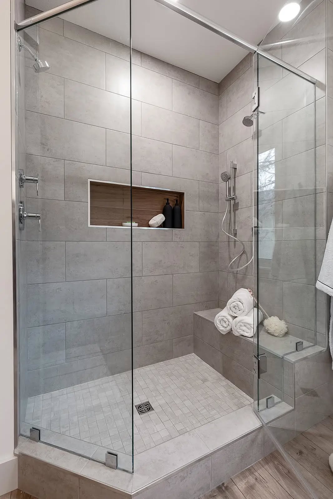 Modern shower with gray tile walls and glass doors. Features a built-in shelf with toiletries and a bench holding rolled towels, creating a clean and relaxing vibe.