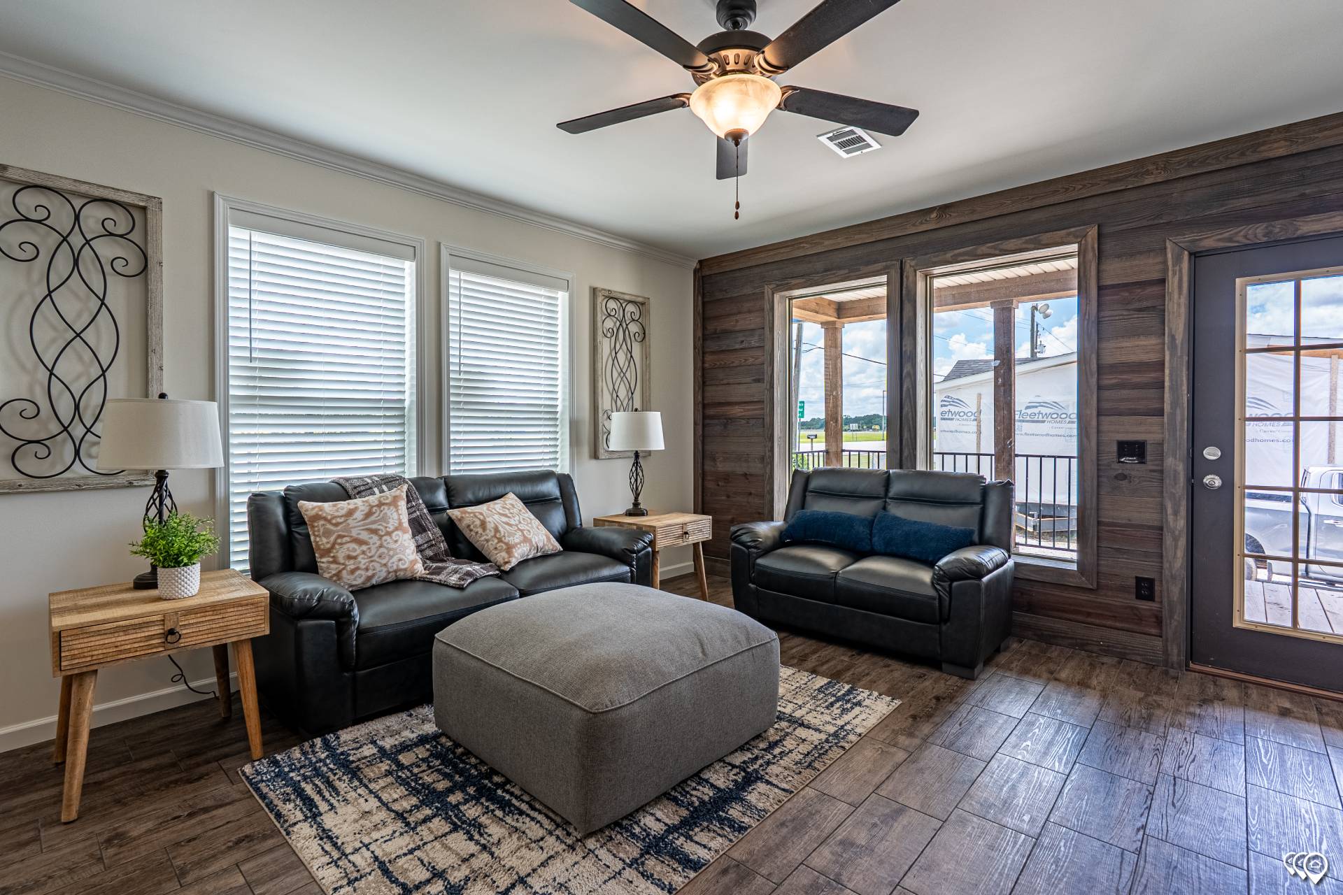 Modern living room with dark sofas, a large grey ottoman, and wooden side tables. Bright windows and a glass door reveal a sunny patio outside.