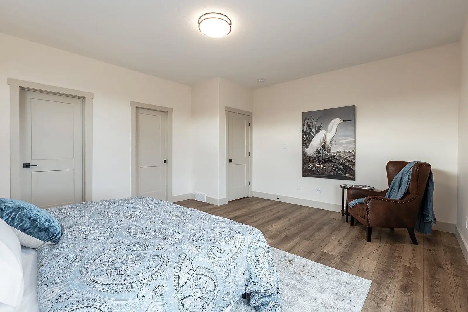 A minimalist bedroom with light walls and wood flooring. A bed with a patterned blue cover, an armchair, a heron painting, and three closed doors are visible. Cozy and serene.