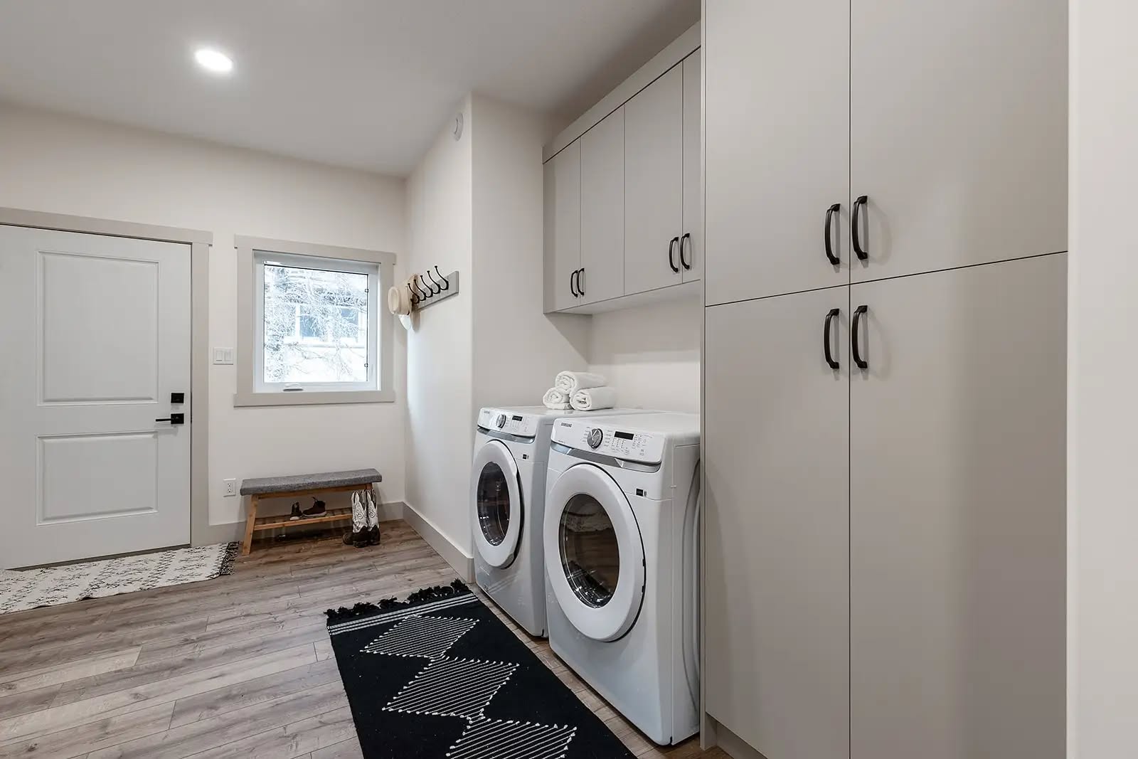 Modern laundry room with white walls, wood floor, washer and dryer, folded towels, black-and-white rug, storage cabinets, and a bench by a window.