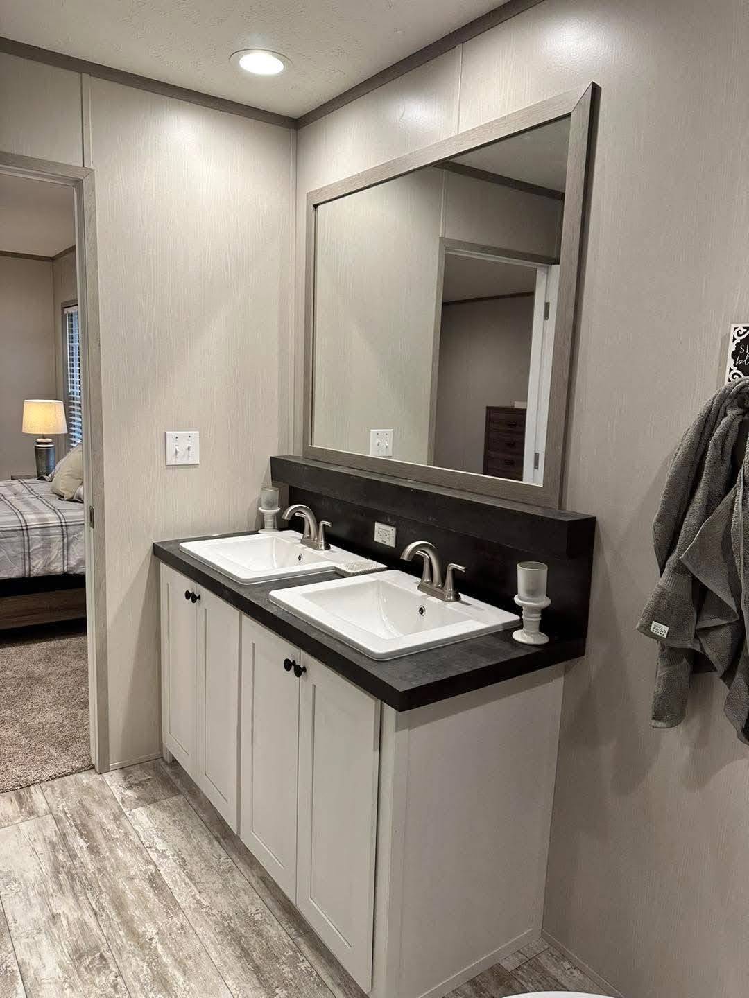 Modern bathroom with dual white sinks on a dark countertop, large mirror, and towel hanging on wall. Bedroom with lamp is visible through the open door.