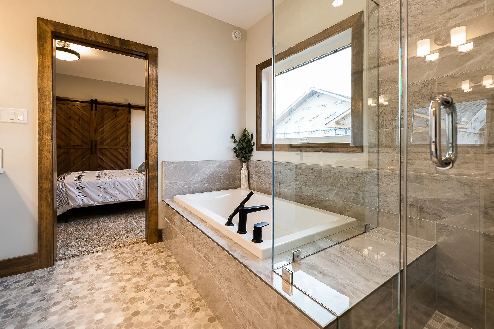 Modern bathroom with a large soaking tub, glass shower, and wooden accents. A doorway reveals a bedroom with a rustic sliding barn door, creating a cozy, elegant atmosphere.