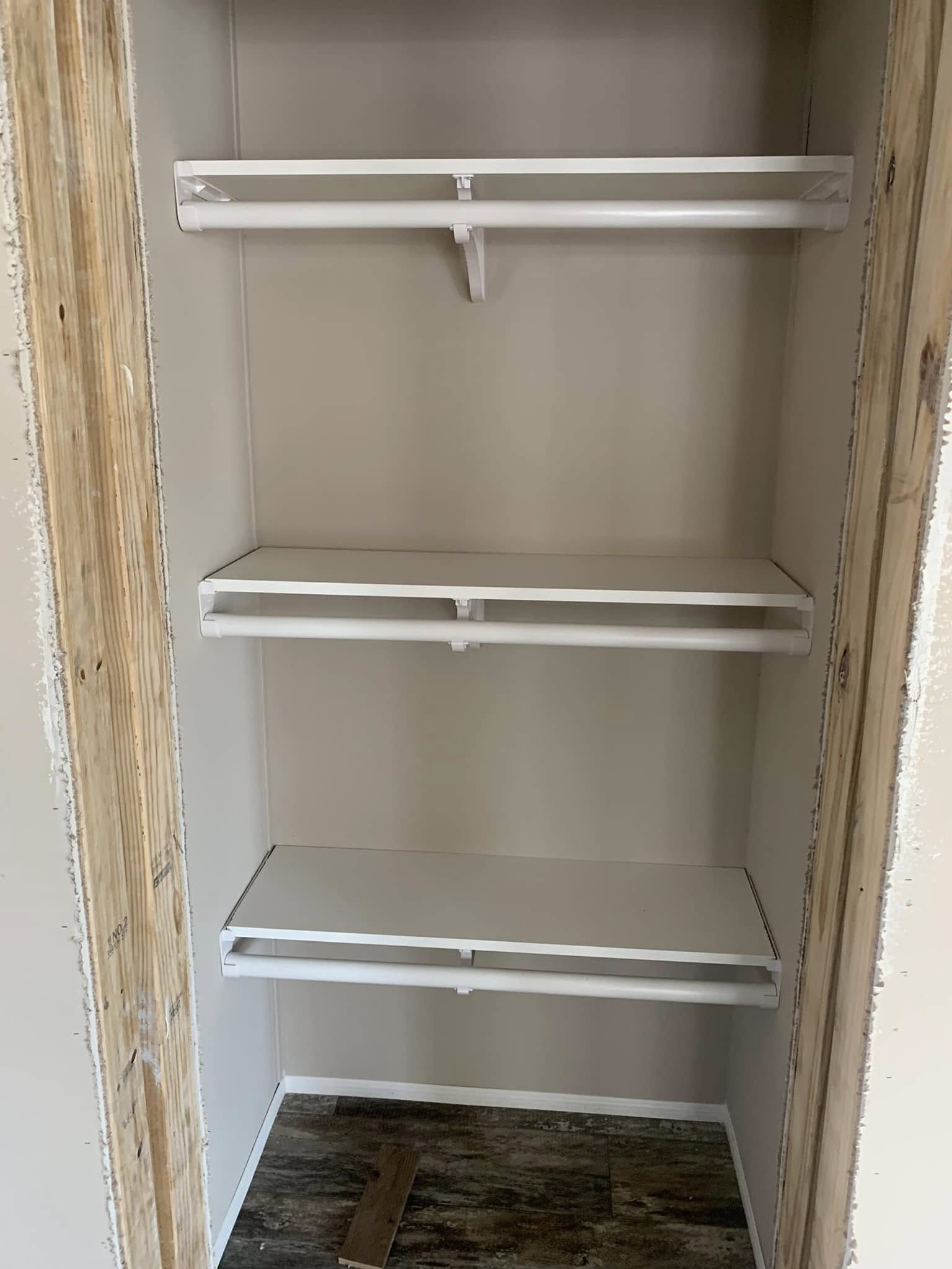 Narrow closet with three white shelves, each featuring a railing below. The wooden frame is unfinished, and the floor has a wood-like pattern.