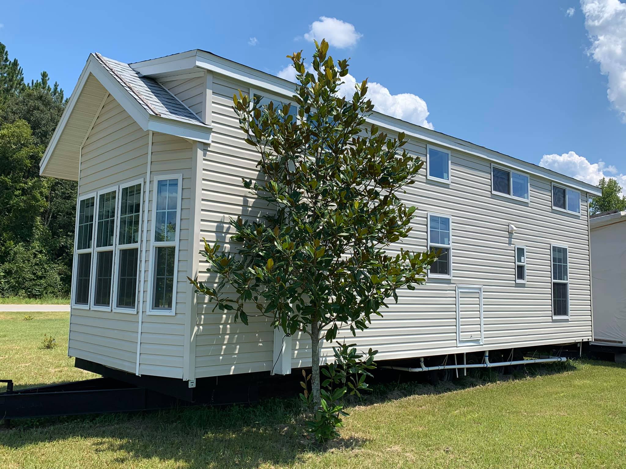 Small beige manufactured home with large windows, set on grassy land. A young tree is in the foreground, and the sky is clear and sunny.