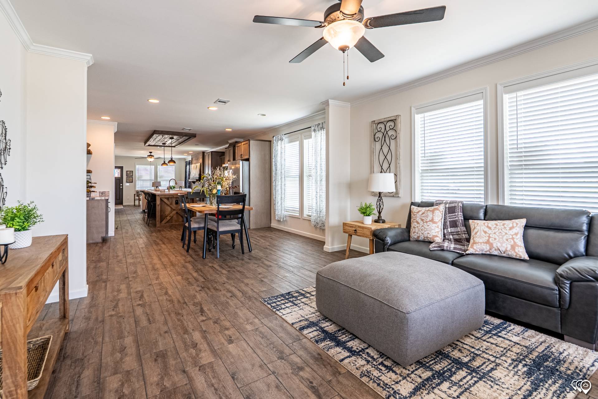 Open-concept living room with wooden floors, featuring a gray sectional sofa, ottoman, and blue rug. A ceiling fan above. Dining area and kitchen visible in background.