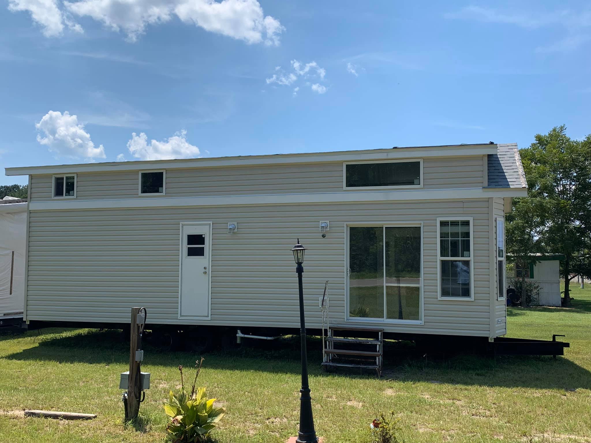 A beige, mobile tiny house with large windows stands on a grassy field under a clear blue sky. A single lamp post is in front, creating a peaceful, minimalist vibe.