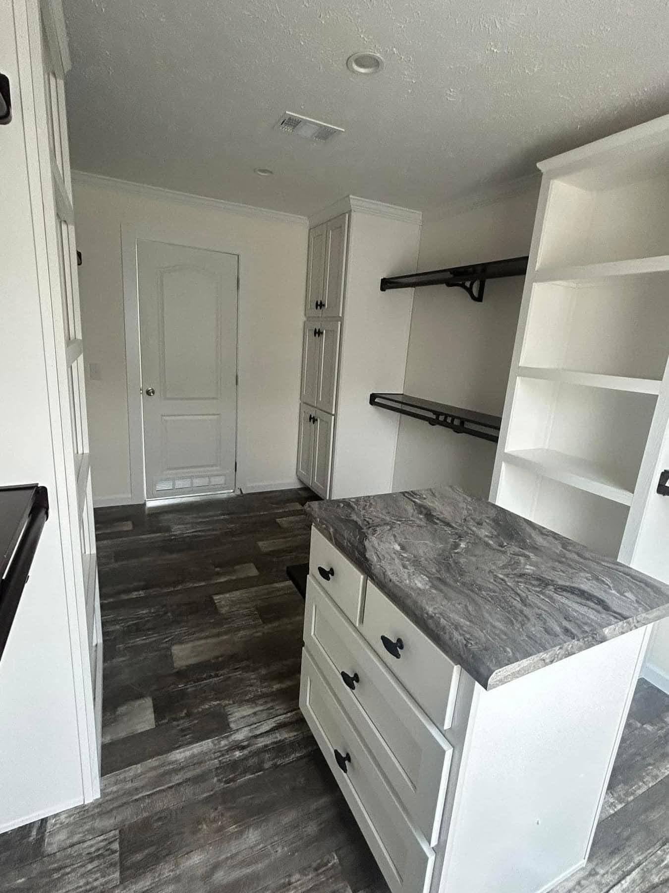 A modern walk-in closet with dark wood flooring, white cabinets, and shelves. A marble-topped island is centered, with a serene, organized feel.