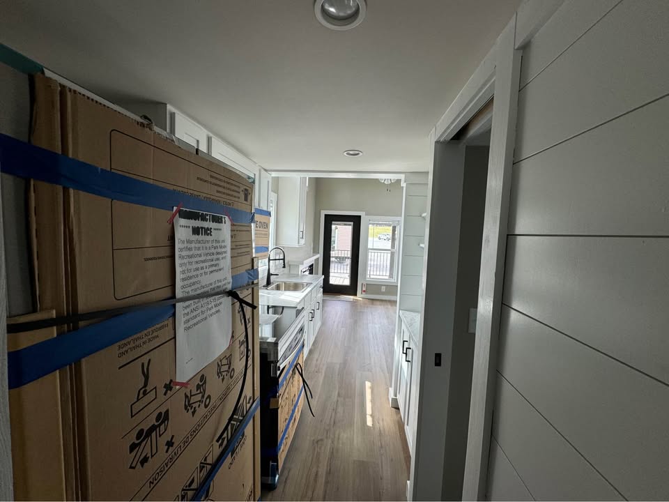 Narrow kitchen under renovation with white cabinets, light wooden flooring, and a black door leading outside. Cardboard boxes and instructions are visible.