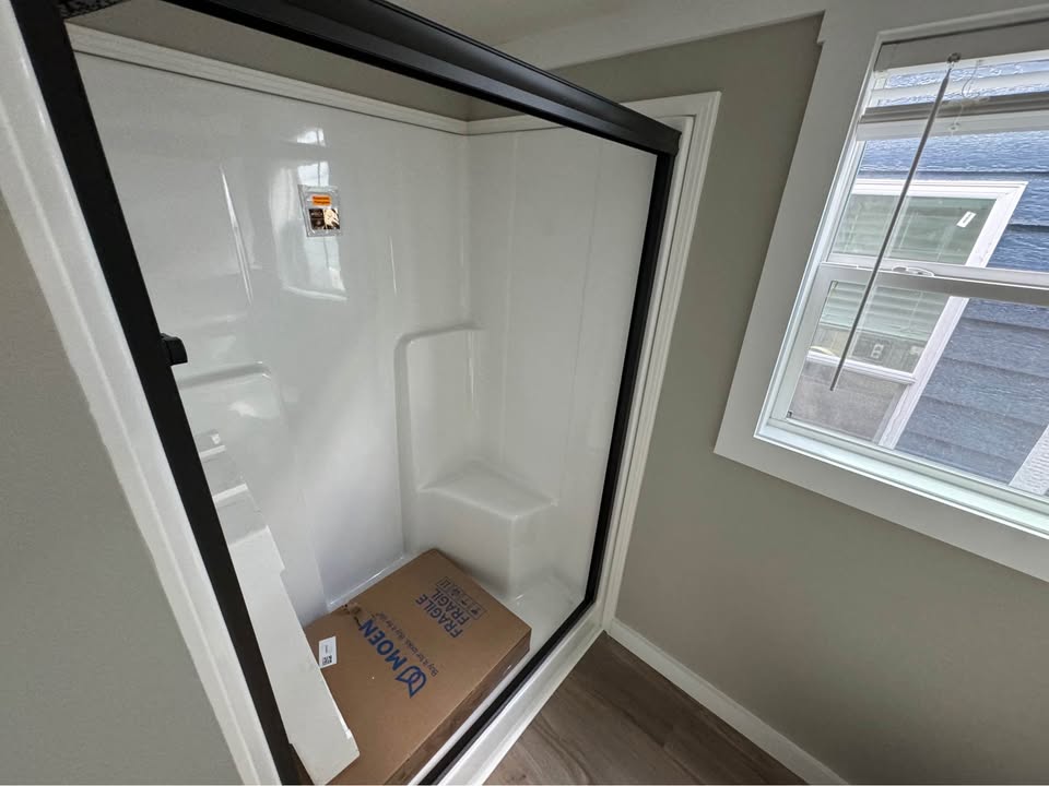 A bathroom with a sleek white shower featuring a black-framed glass door. There's an unopened Moen box inside the shower, and a sunlit window is nearby.