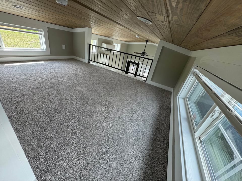 Cozy attic space with gray carpet and wood-paneled ceiling. Three windows provide natural light. A black railing overlooks the lower level. Serene ambiance.