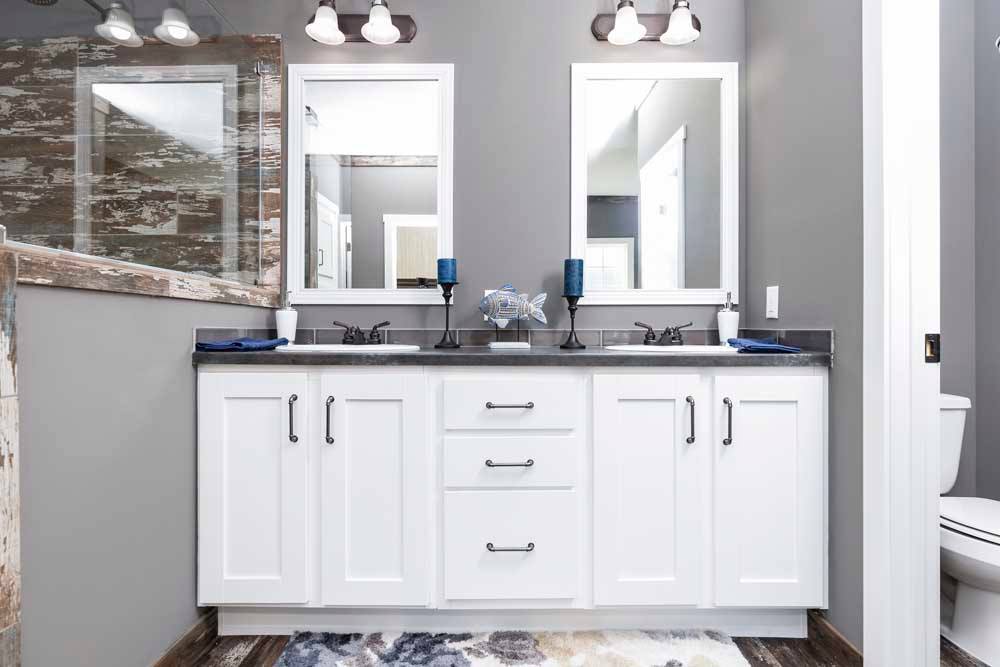 Modern bathroom with dual sinks, white cabinets, and gray walls. Two mirrors above sinks, blue candles, decorative fish, and shower in view. Calm atmosphere.