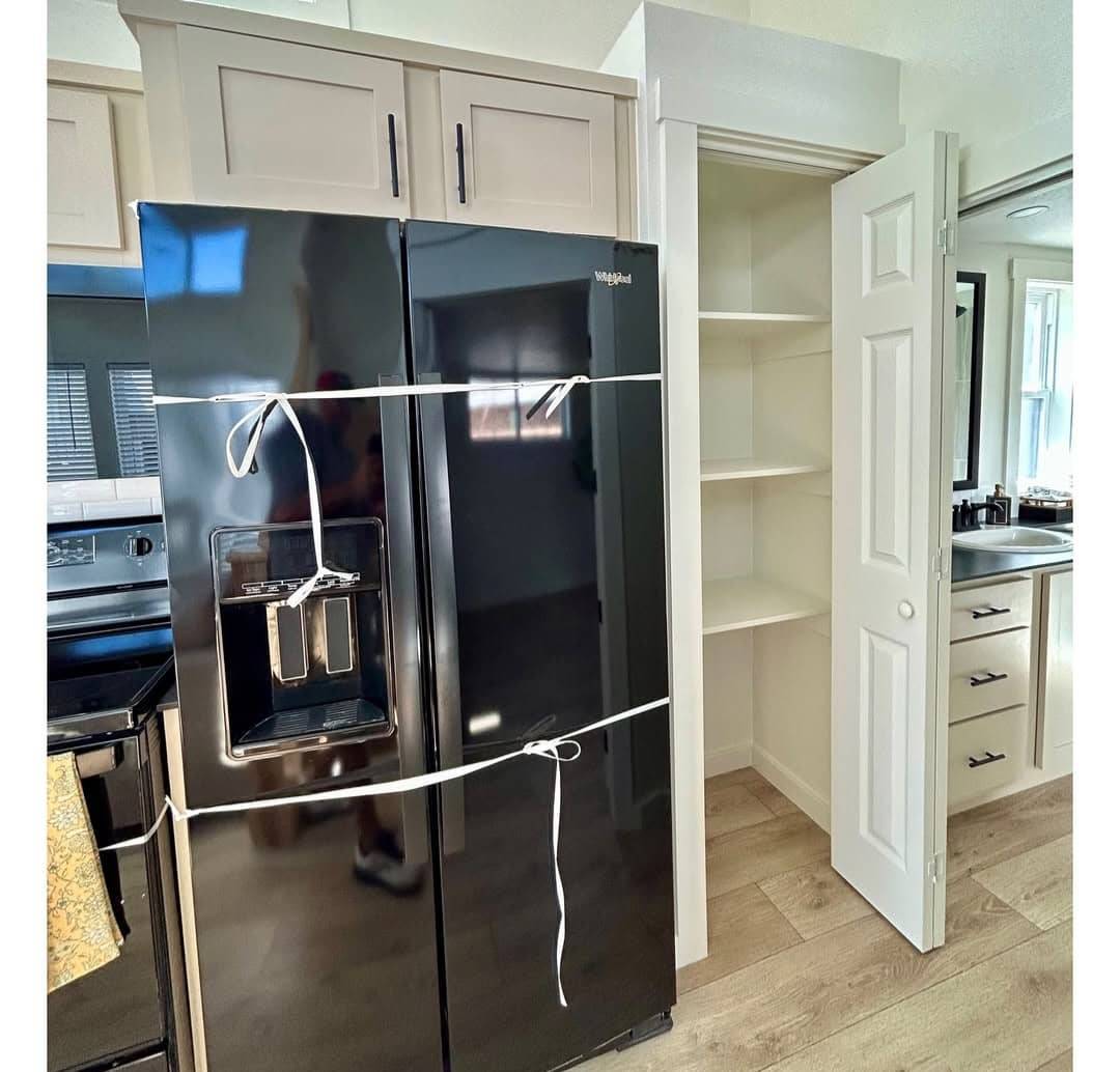 Black refrigerator secured with white cords beside an open pantry door in a modern kitchen. Light wooden flooring adds a warm contrast.