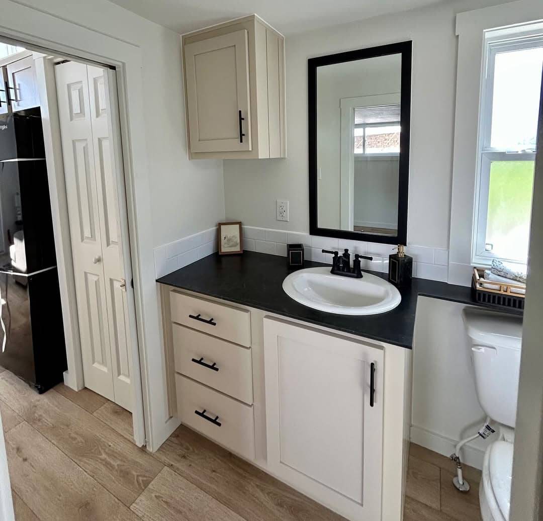 Small bathroom with light wood flooring, white cabinets, and black countertop. Features a white sink, black faucet, and rectangular mirror.
