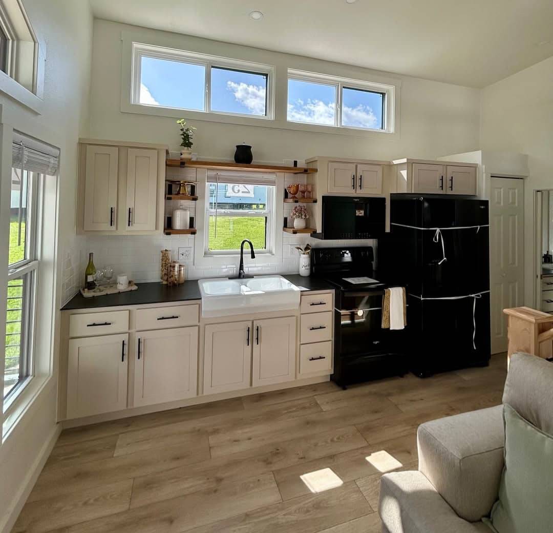 Modern kitchen with light wood cabinets, black countertops, and appliances. Large windows let in natural light, creating a bright, inviting space.