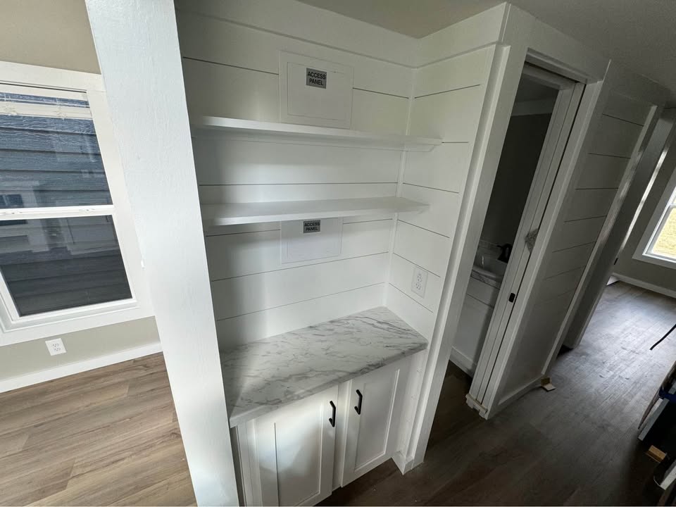 White hallway nook with marble countertop, two open shelves, and small cabinet, near doorways and a window on wood flooring, conveying minimalist style.