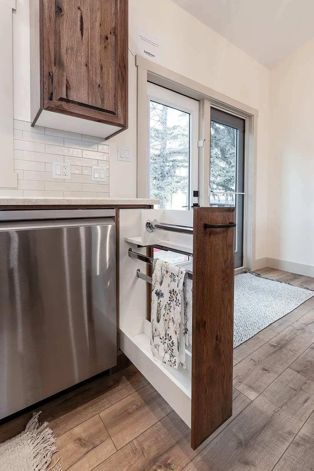 A modern kitchen with a pull-out towel rack inside a wooden cabinet, stainless steel dishwasher, and sliding glass door, creating a fresh, organized look.