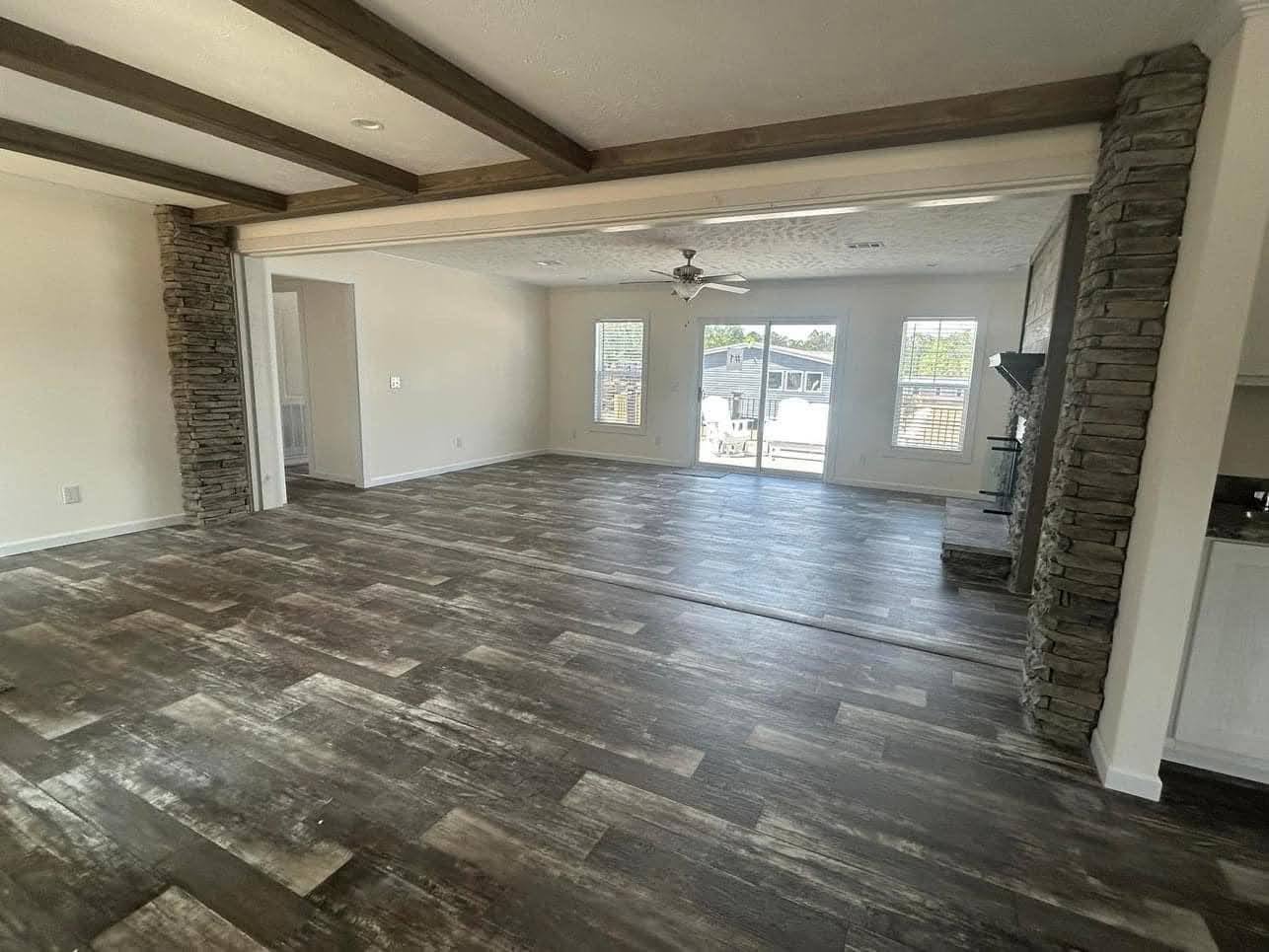 Spacious living room with rustic wooden beams and weathered wood flooring. Stone wall accents and a ceiling fan add a cozy, inviting touch.