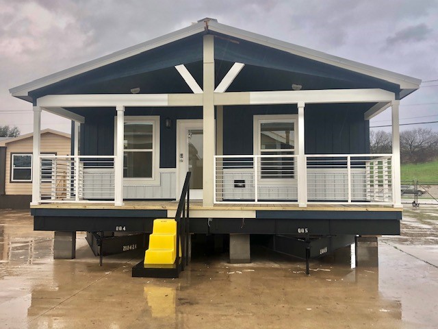 Modern prefabricated home with a dark exterior and white trim. Features a covered porch with railings, yellow ramp, and concrete surroundings on a rainy day.