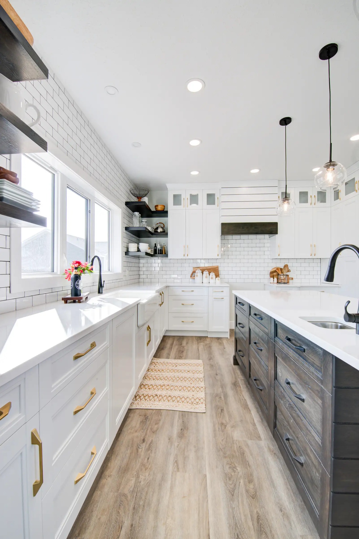 Bright modern kitchen with white cabinets, brass handles, and a wooden floor. Features black and white counters, pendant lights, and a farmhouse sink.