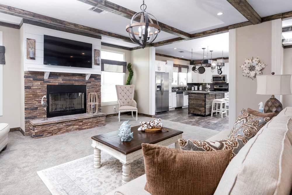 Elegant living room with a cozy fireplace, beige sofa, decorative pillows, and wooden coffee table. Open view to a modern kitchen with stainless appliances.