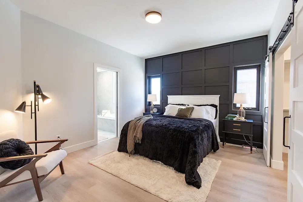 Modern bedroom with dark paneled feature wall, king-sized bed with plush black bedding and white headboard. Soft lighting, cozy armchair, and wood flooring.