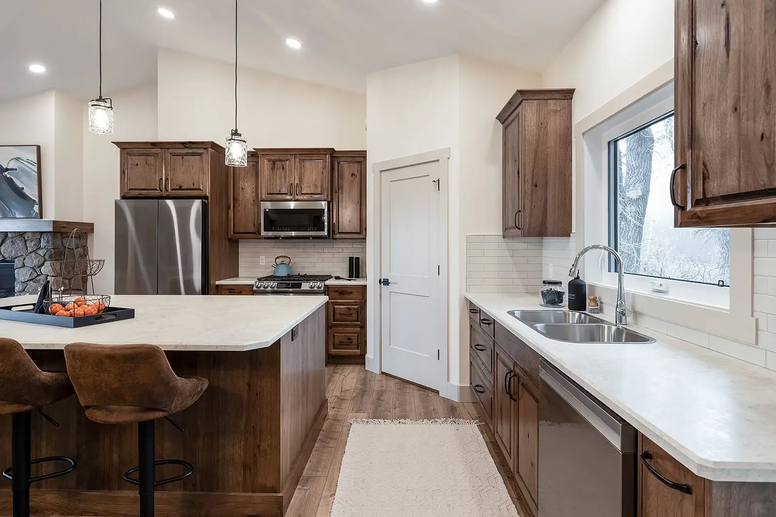 Rustic kitchen with wooden cabinetry, stainless steel appliances, and a central island with two stools. Bright, cozy ambiance with pendant lighting.