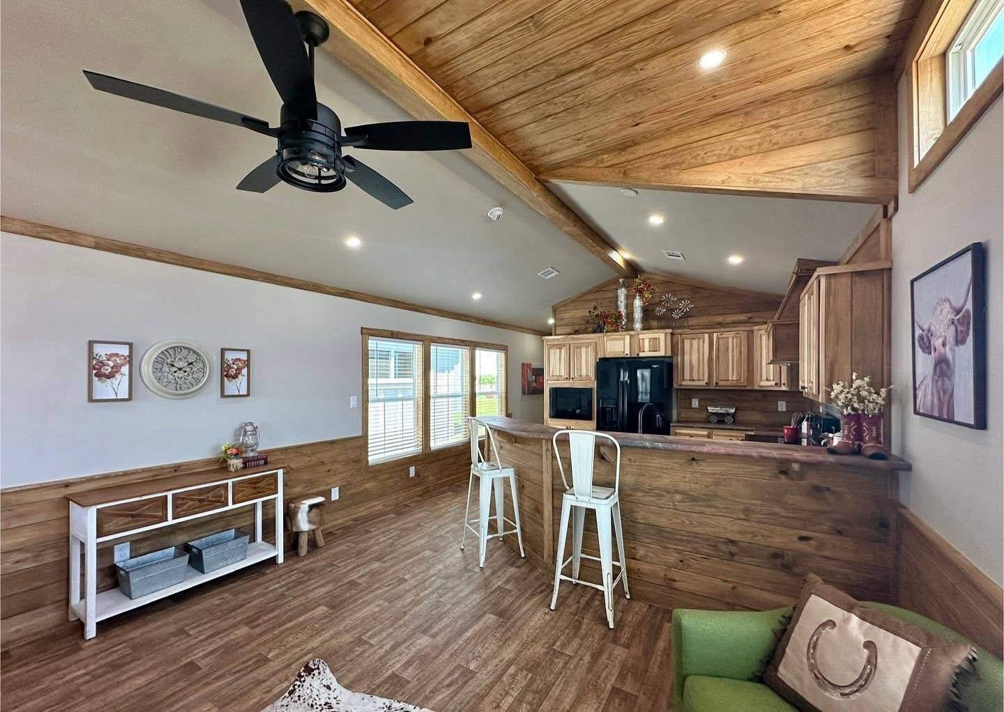 Rustic open-plan living area with wooden accents. Features a kitchen with bar stools, ceiling fan, green chair, wall clock, and bright windows. Cozy and inviting.